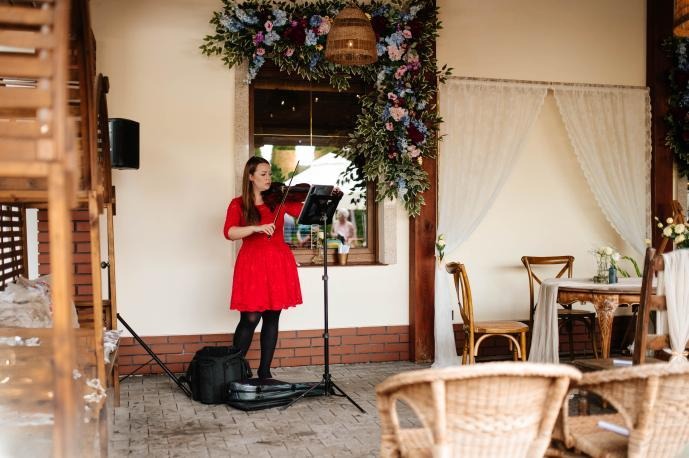 Skrzypaczka w czerwonej sukience gra na skrzypcach na tle dekoracji kwiatowych i białych zasłon, tworząc elegancką oprawę muzyczną wydarzenia.