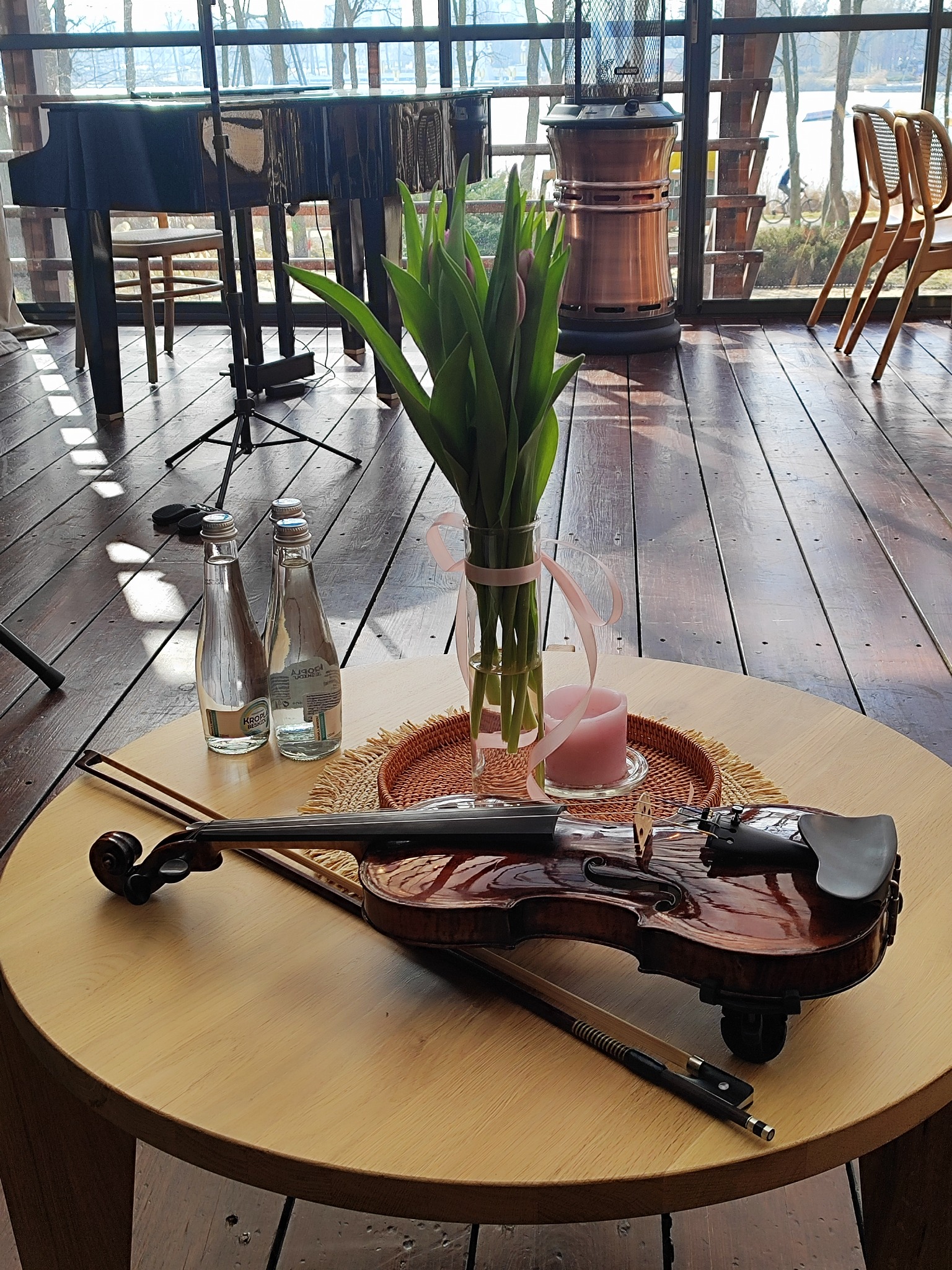 Stylowe wnętrze z pianinem w tle, na okrągłym stole skrzypce, smyczek, wazon z tulipanami i świeca. Dwie butelki wody mineralnej. Drewniana podłoga i duże okna.