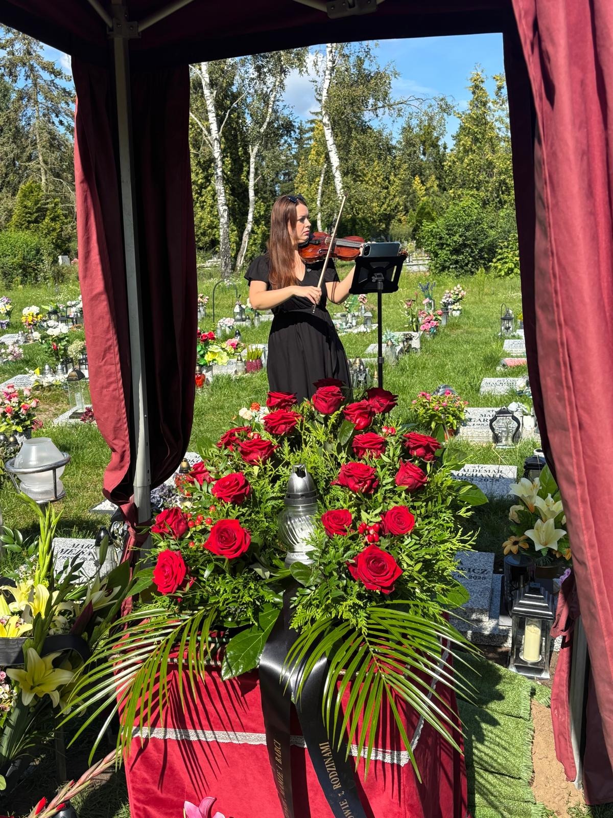 Skrzypaczka w czarnej sukience gra na cmentarzu, na tle nagrobków i zieleni, z bukietem czerwonych róż na pierwszym planie. Uroczystość pogrzebowa.