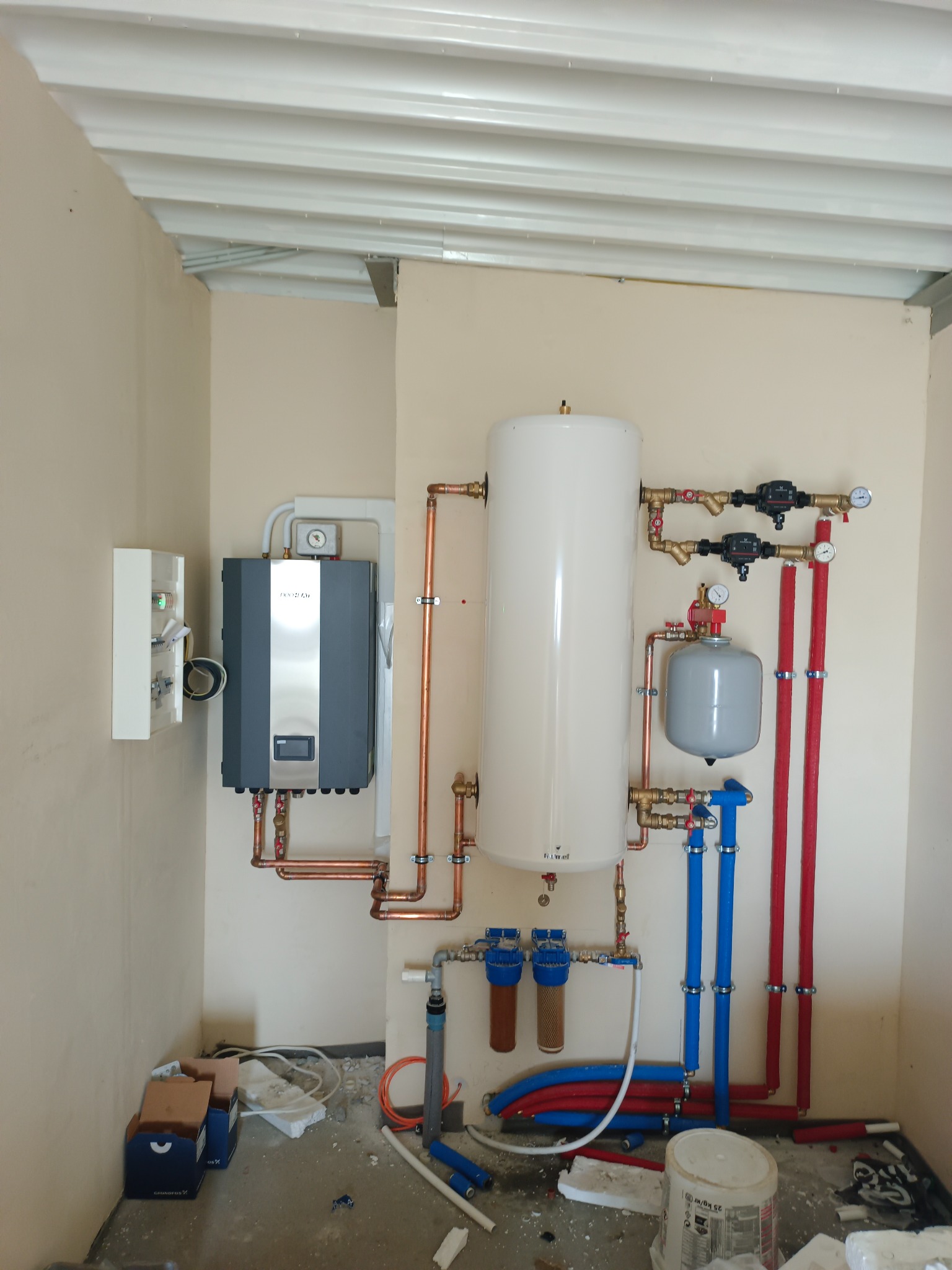 Nowoczesna instalacja CO z zasobnikiem i filtrami, rury miedziane i izolowane, zamontowana w pomieszczeniu z surowym wykończeniem. Widoczne elementy sterujące i zabezpieczające.