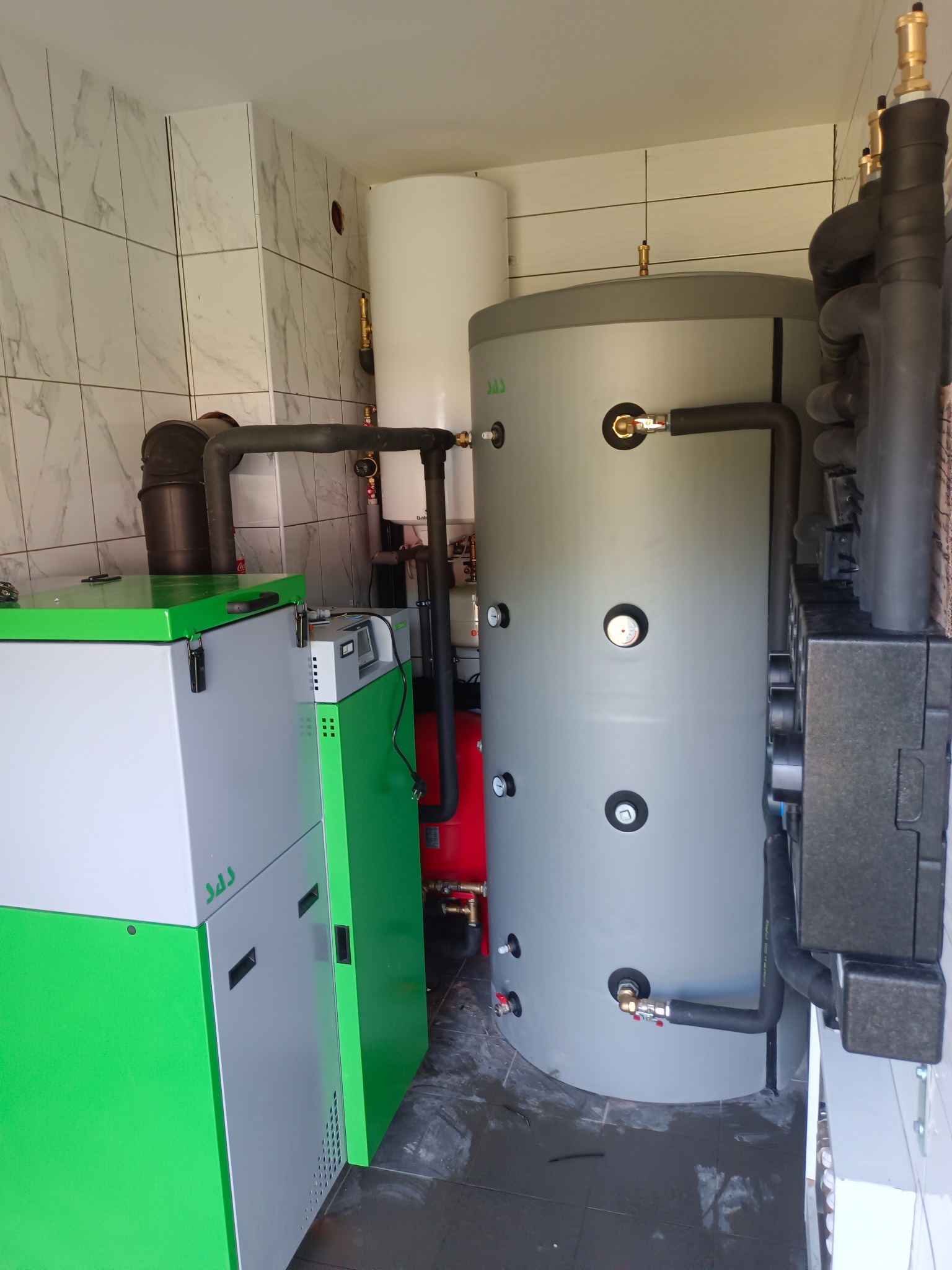 Kotłownia z kotłem SAS, zbiornikiem akumulacyjnym i instalacją. Białe płytki na ścianach. Kompaktowe rozwiązanie grzewcze w małym pomieszczeniu.