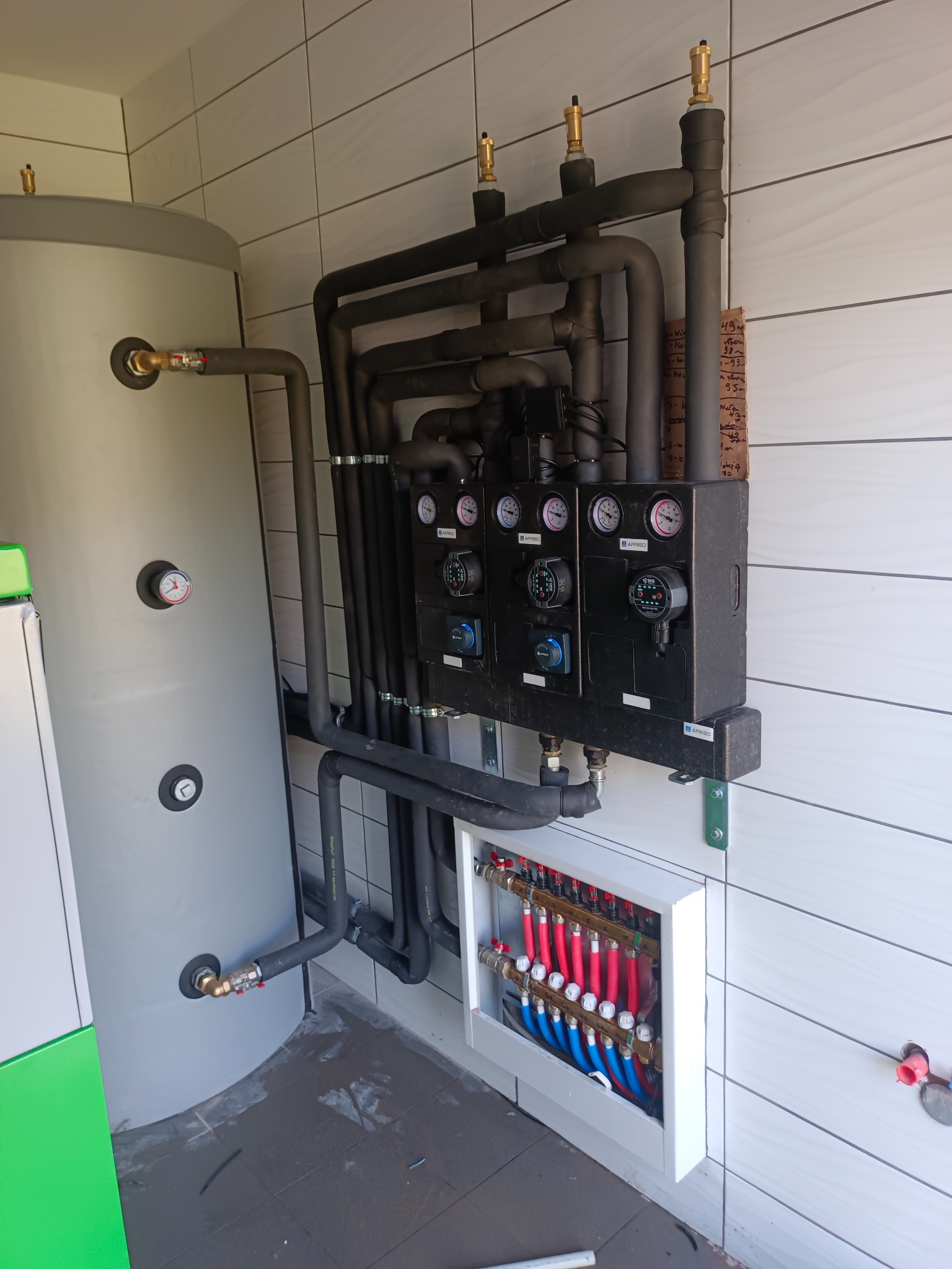 Kompletna instalacja ogrzewania podłogowego z rozdzielaczem, pompami obiegowymi i zbiornikiem buforowym. Widoczne rury w kolorach czerwonym i niebieskim.