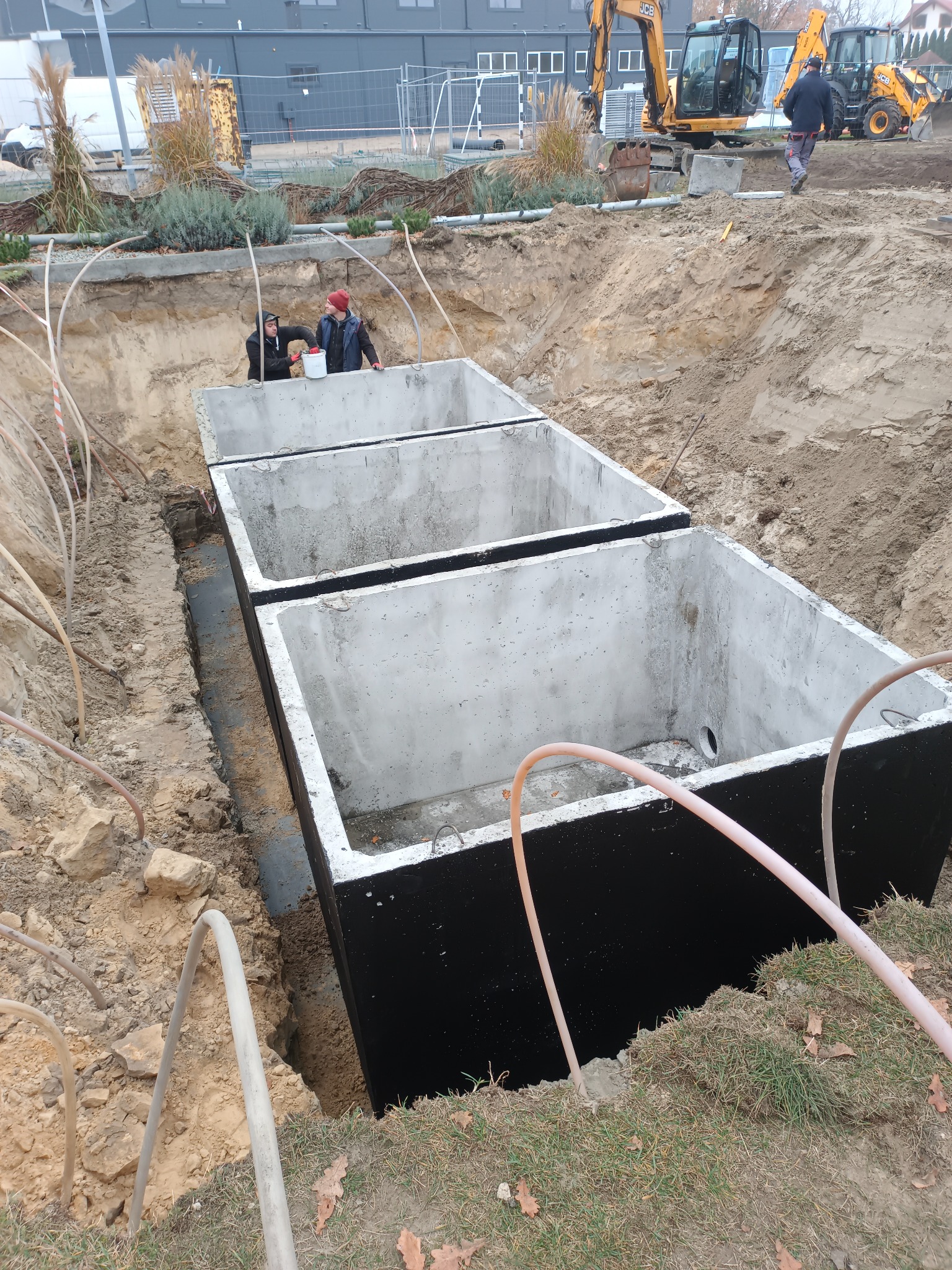 Montaż betonowych zbiorników w wykopie, prawdopodobnie szamba lub oczyszczalni. Widoczni pracownicy i sprzęt budowlany w tle. Ujęcie z góry, podkreślające głębokość wykopu.