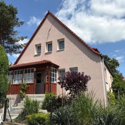 ELEVARO - Jasnoróżowy dom jednorodzinny z czerwonym dachem i drewnianą werandą, otoczony zielenią i niskim ogrodzeniem. Błękitne niebo z chmurami w tle.