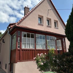 ELEVARO - Jasnoróżowy dom z czerwonym dachem i drewnianą werandą, numer 29 na ścianie. W tle błękitne niebo z chmurami. Z przodu krzew róż i zielony kosz na śmieci.