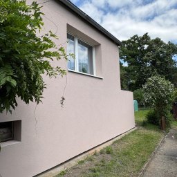 ELEVARO - Jasnoróżowa elewacja domu jednorodzinnego z białymi oknami, widok z boku, zieleń w tle i na pierwszym planie. Świeżo położony tynk.