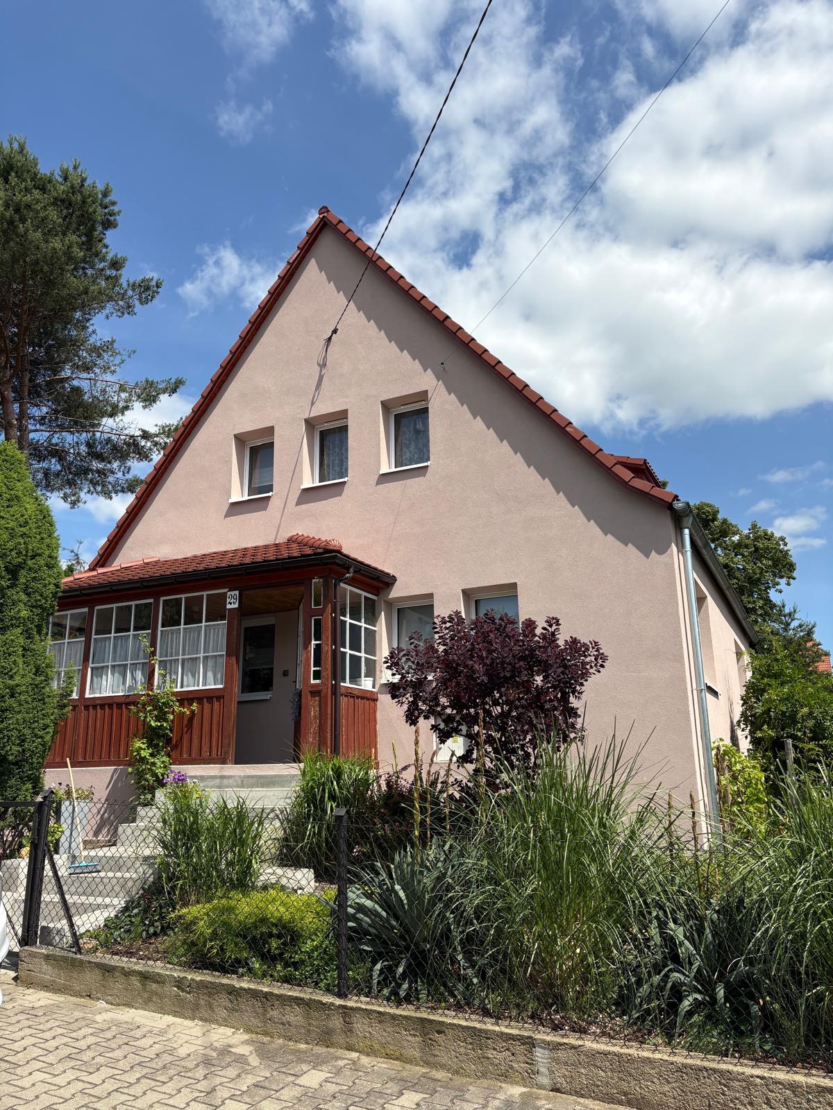 Jasnoróżowy dom jednorodzinny z czerwonym dachem i drewnianą werandą, otoczony zielenią i niskim ogrodzeniem. Błękitne niebo z chmurami w tle.