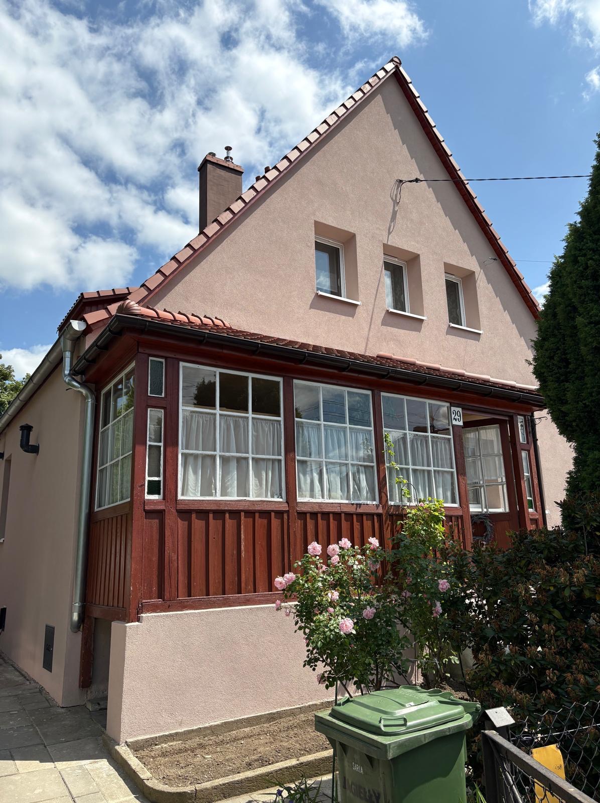 Jasnoróżowy dom z czerwonym dachem i drewnianą werandą, numer 29 na ścianie. W tle błękitne niebo z chmurami. Z przodu krzew róż i zielony kosz na śmieci.