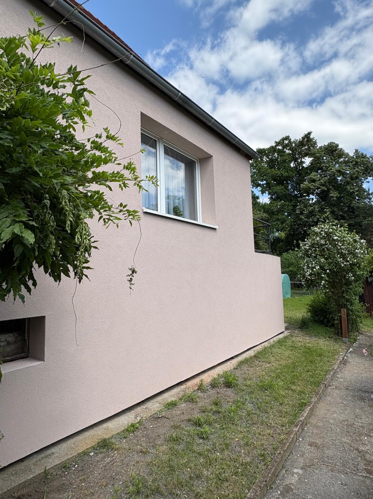Jasnoróżowa elewacja domu jednorodzinnego z białymi oknami, widok z boku, zieleń w tle i na pierwszym planie. Świeżo położony tynk.