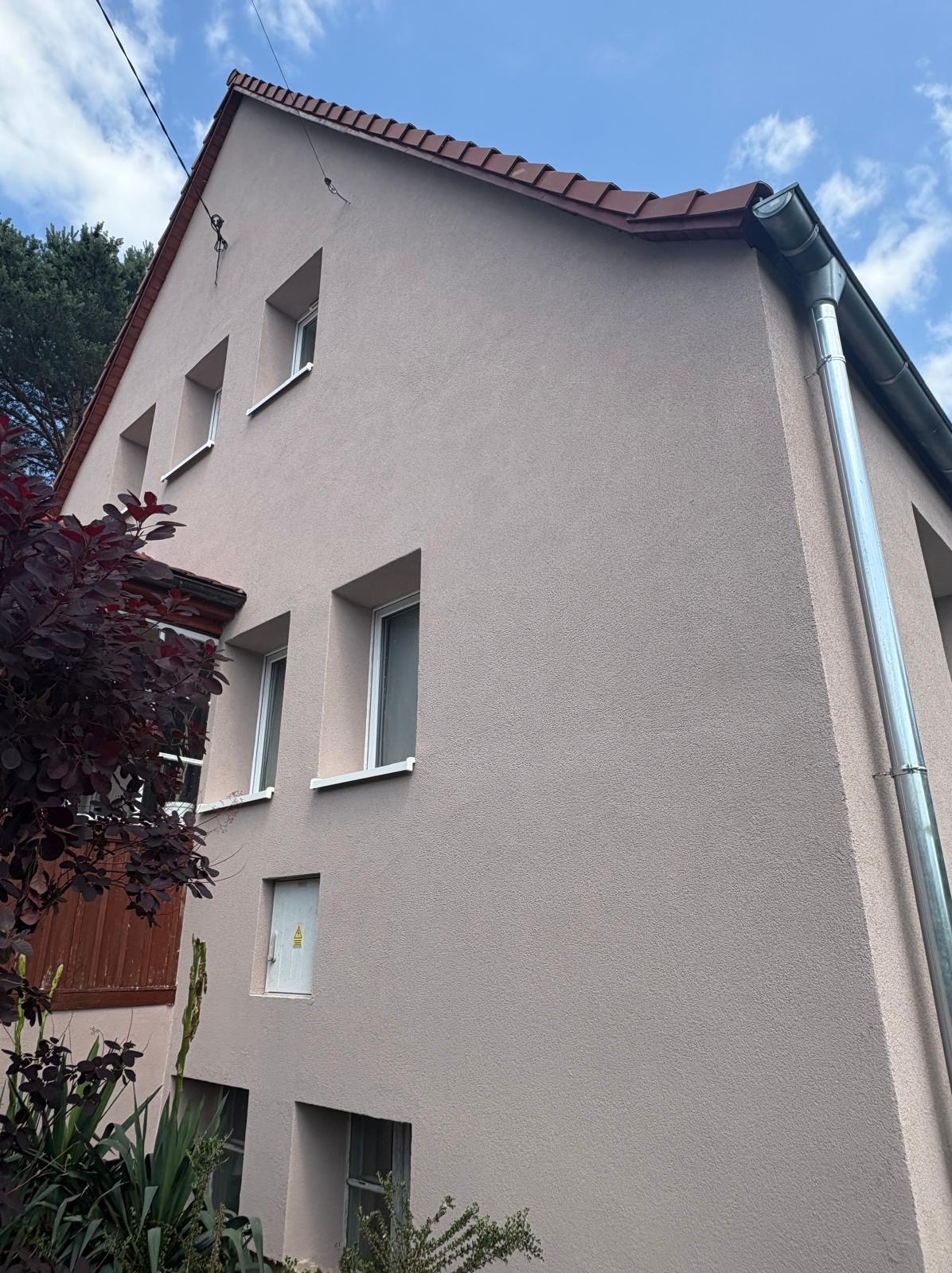 Elewacja domu jednorodzinnego w odcieniach beżu, z widocznymi oknami, rynną i fragmentem dachu. Ujęcie z dołu, podkreślające strukturę tynku.