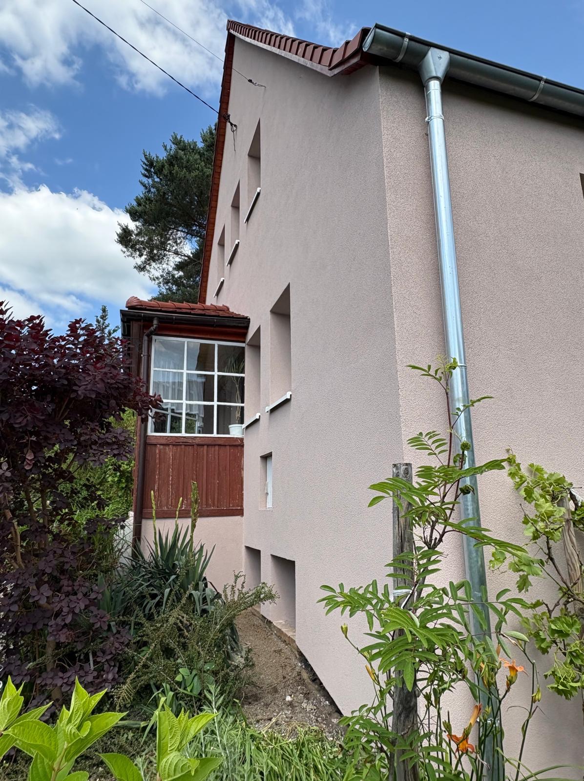 Jasnoróżowa elewacja domu z drewnianym balkonem i metalową rynną, otoczona zielenią. Widok z dołu, podkreślający strukturę tynku i detale architektoniczne.