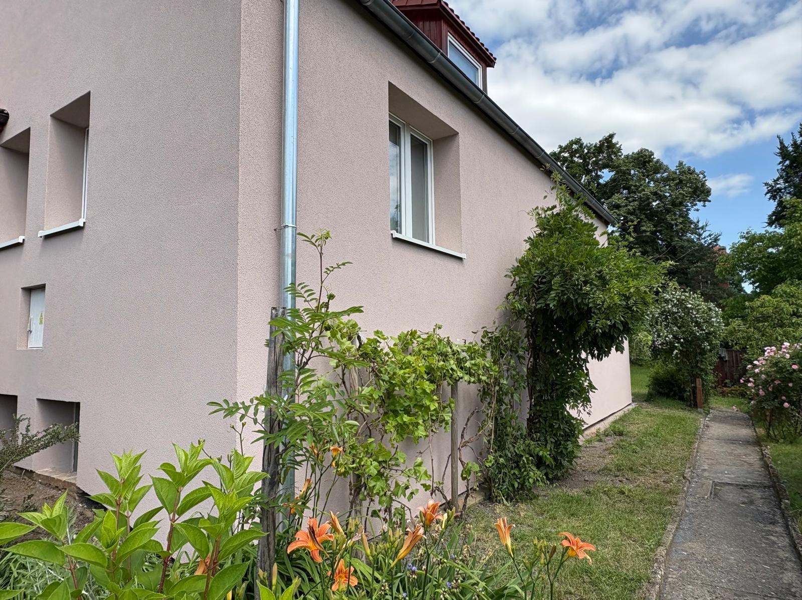Ocieplona elewacja domu w odcieniach beżu, z widoczną rynną i roślinnością przy ścianie. Świeżo pomalowana fasada z oknami i fragmentem dachu.