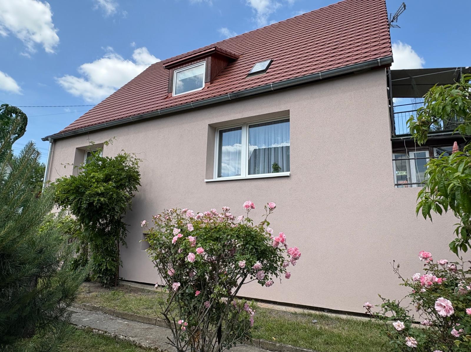 Dom jednorodzinny z jasną elewacją i czerwoną dachówką, otoczony zielenią i różowymi krzewami róż. Widoczne okna z białymi ramami i fragment nieba z chmurami.