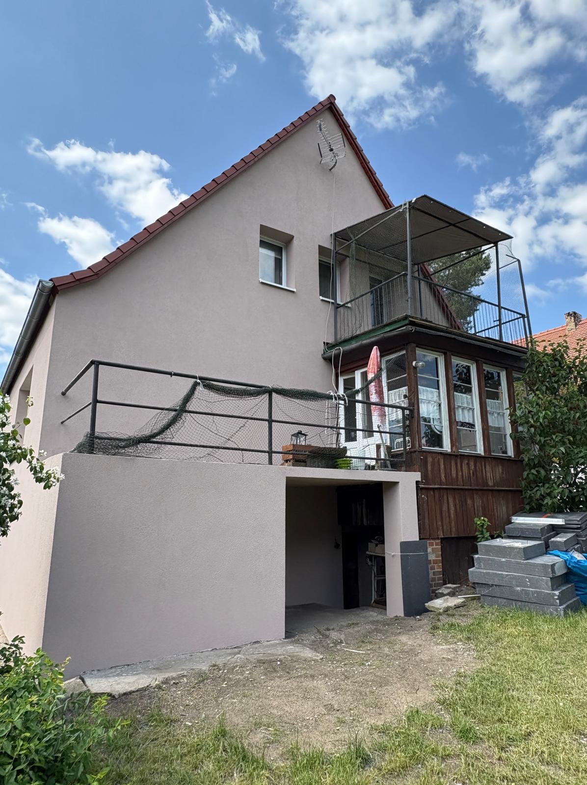 Dom z ocieploną elewacją w odcieniach beżu, z balkonem i zadaszeniem. Widoczne elementy wykończeniowe i otoczenie budynku.