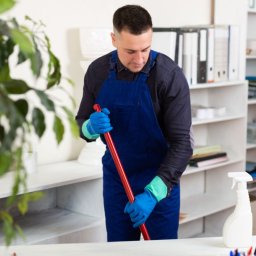 Sergii Palanskyi Działalność Gospodarcza - Pracownik w niebieskim kombinezonie myje podłogę w biurze, używając mopa z czerwoną rączką. W tle regał z segregatorami i biurko z lampką oraz rośliny doniczkowe.