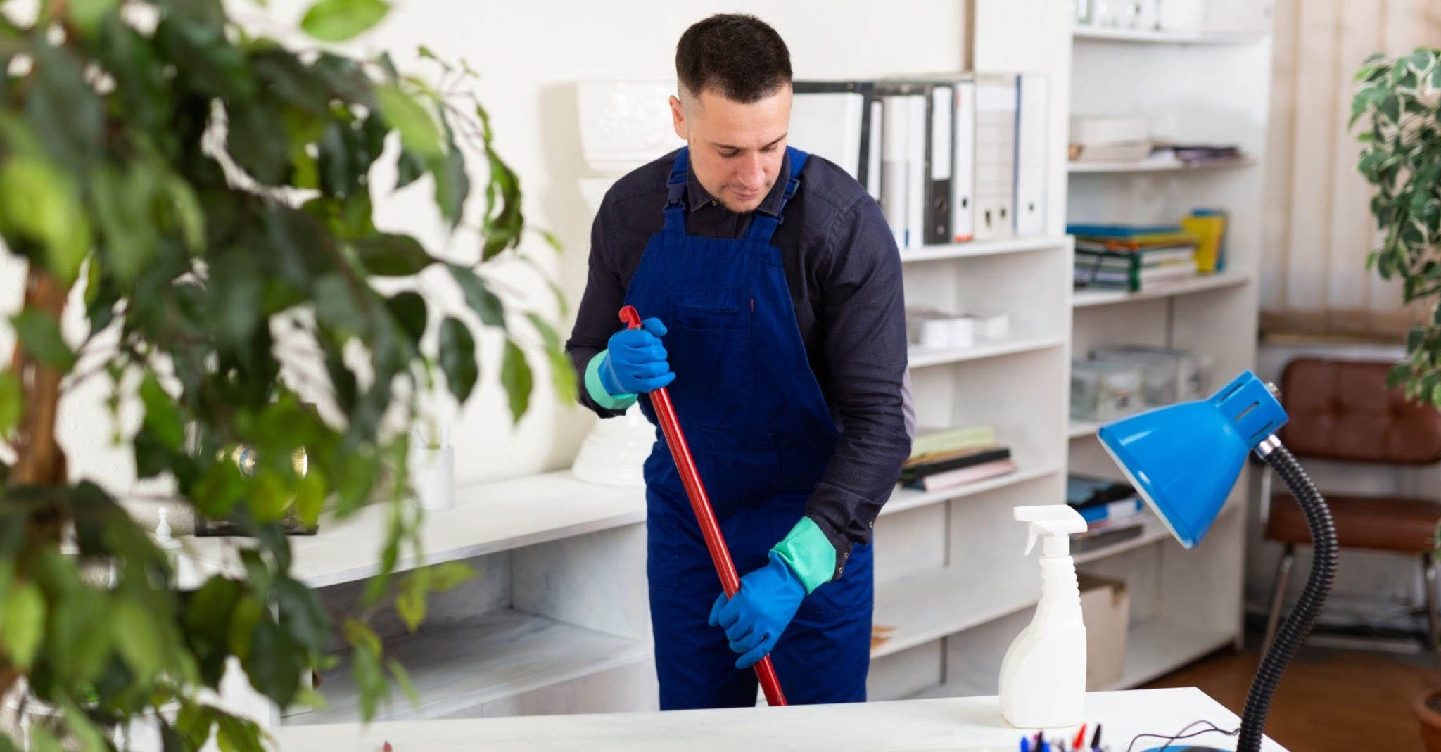 Pracownik w niebieskim kombinezonie myje podłogę w biurze, używając mopa z czerwoną rączką. W tle regał z segregatorami i biurko z lampką oraz rośliny doniczkowe.