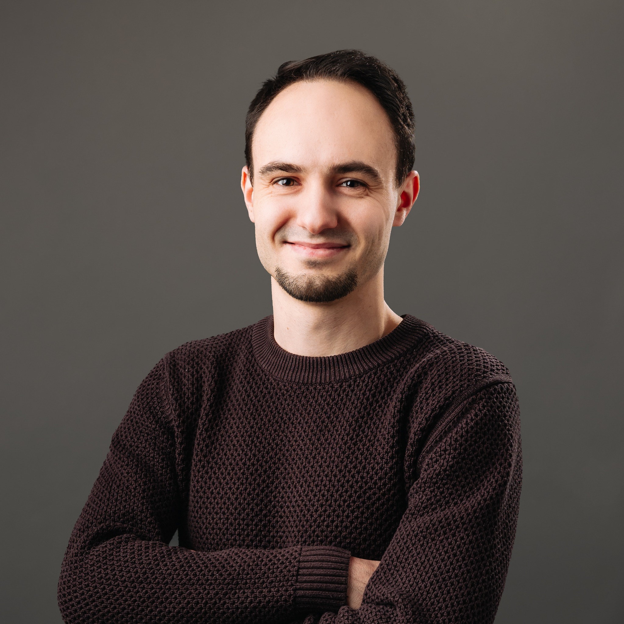 Uśmiechnięty programista w swetrze, z brodą, na szarym tle, prezentuje się profesjonalnie z założonymi rękami. Portret.