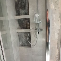 B.O.S_BuD - Nowoczesna kabina prysznicowa z szarymi płytkami i wnękami z ciemniejszym akcentem. Chromowana armatura i szklane drzwi. Widoczna dłoń otwierająca kabinę.