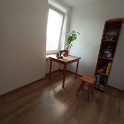 B.O.S_BuD - Pokój z drewnianą podłogą, stołem przy oknie, rośliną doniczkową i narzędziami. Widoczny regał z półkami i mały stołek. Przygotowania do pracy.