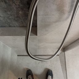 B.O.S_BuD - Nowoczesna kabina prysznicowa z odpływem liniowym i szklanymi drzwiami, wykończona szarą płytką imitującą beton. Widoczne stopy osoby fotografującej.