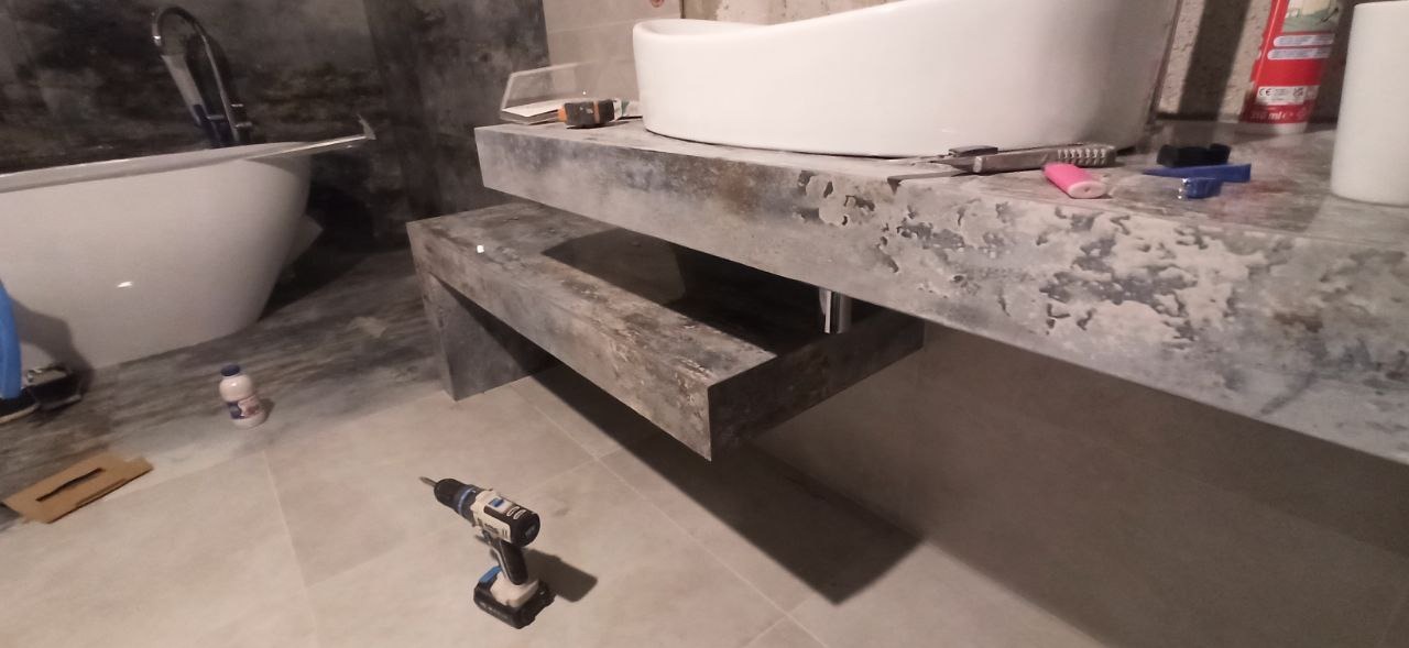 Montaż umywalki nablatowej z blatem imitującym beton. Widoczna wiertarka, wanna i akcesoria. Surowy styl łazienki w trakcie wykańczania.