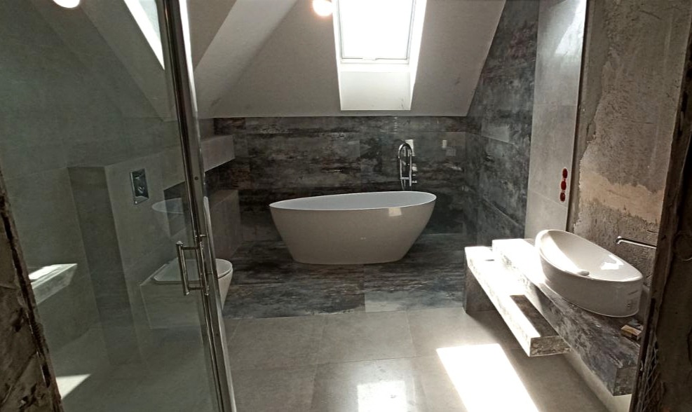 Nowoczesna łazienka na poddaszu z wolnostojącą wanną, umywalką na blacie z kamienia i kabiną prysznicową. Widoczne okno dachowe i szare płytki imitujące kamień.