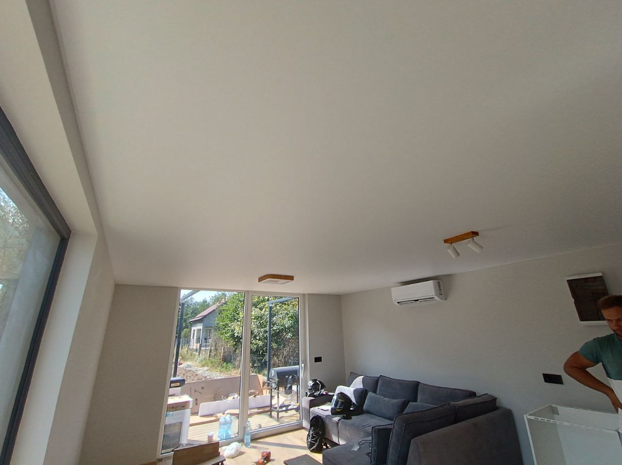 Jasne wnętrze po malowaniu: gładka, biała sufit i ściany, szary narożnik, okno z widokiem na zieleń. Widoczny klimatyzator i lampa sufitowa. Częściowo widoczna osoba.