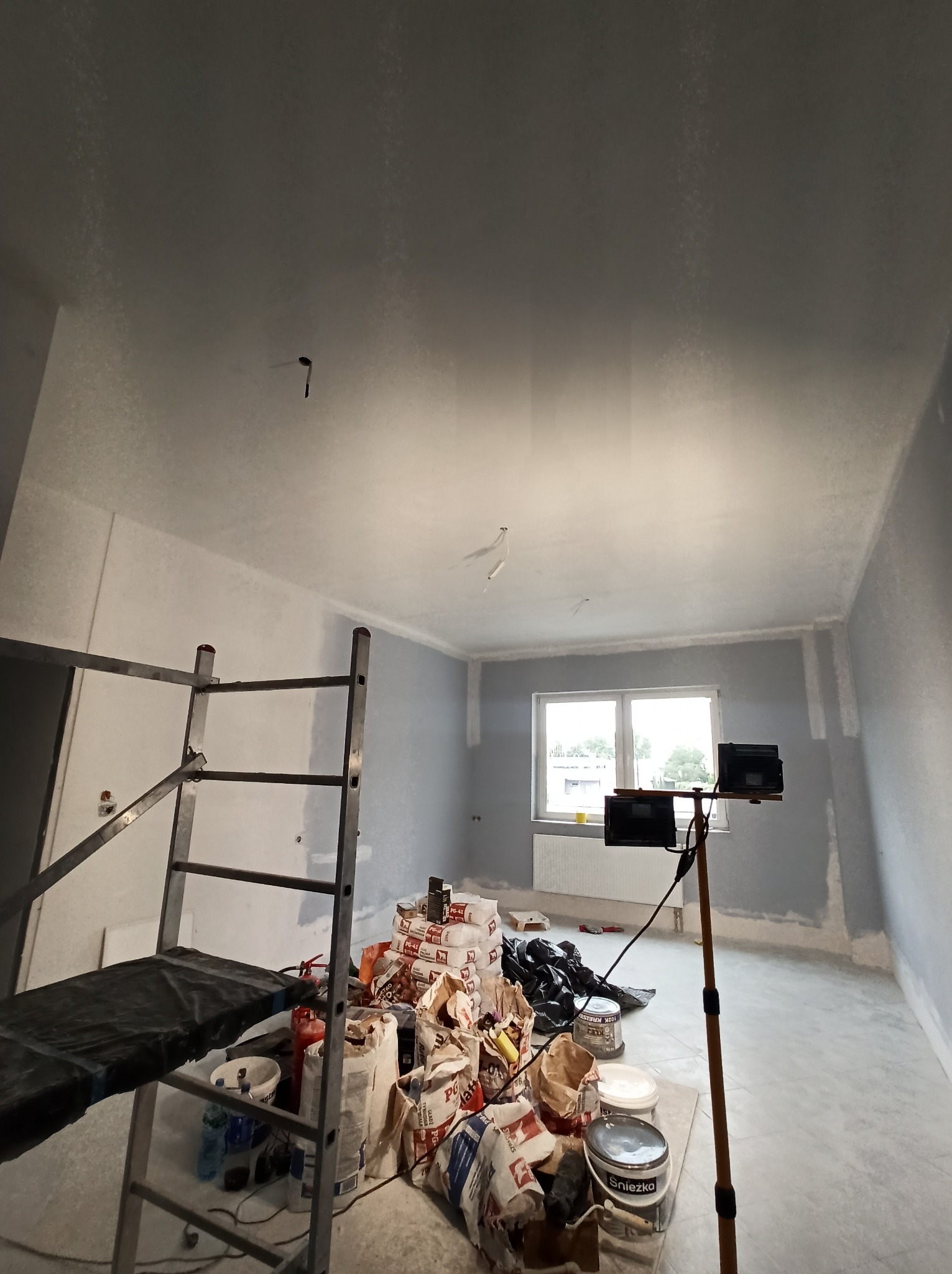 Pokój w trakcie malowania w Krakowie. Widoczne worki z gładzią, drabina malarska i lampa oświetleniowa. Ściany częściowo pomalowane na szaro.