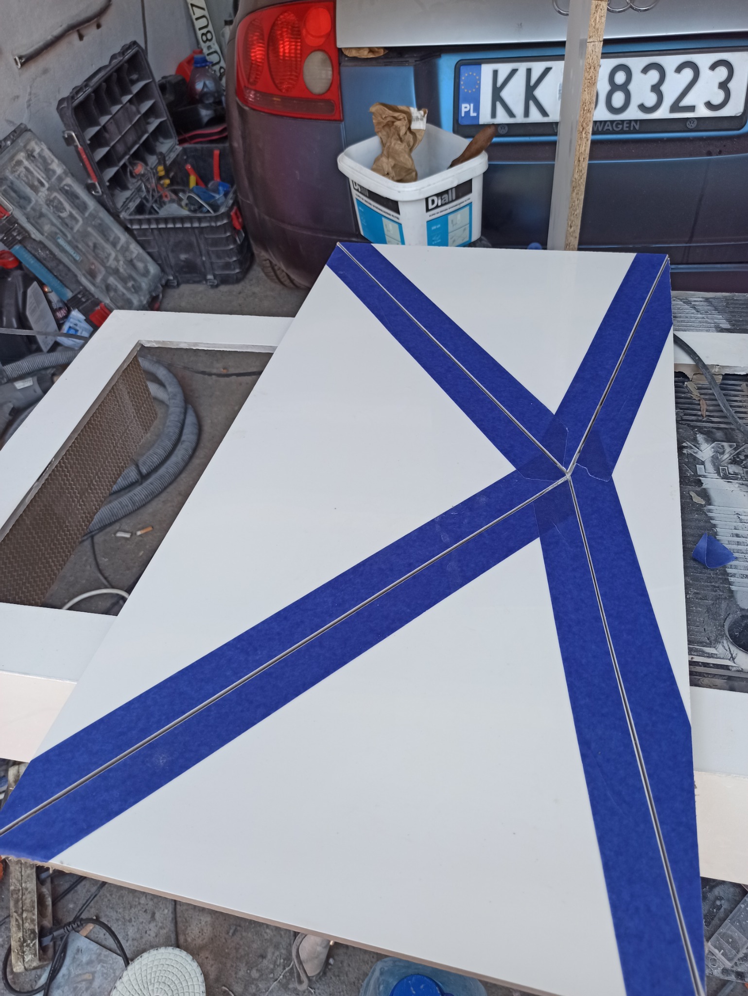 Biała płyta z niebieską taśmą malarską ułożoną w kształt litery X, przygotowana do malowania. W tle widoczne narzędzia i samochód.
