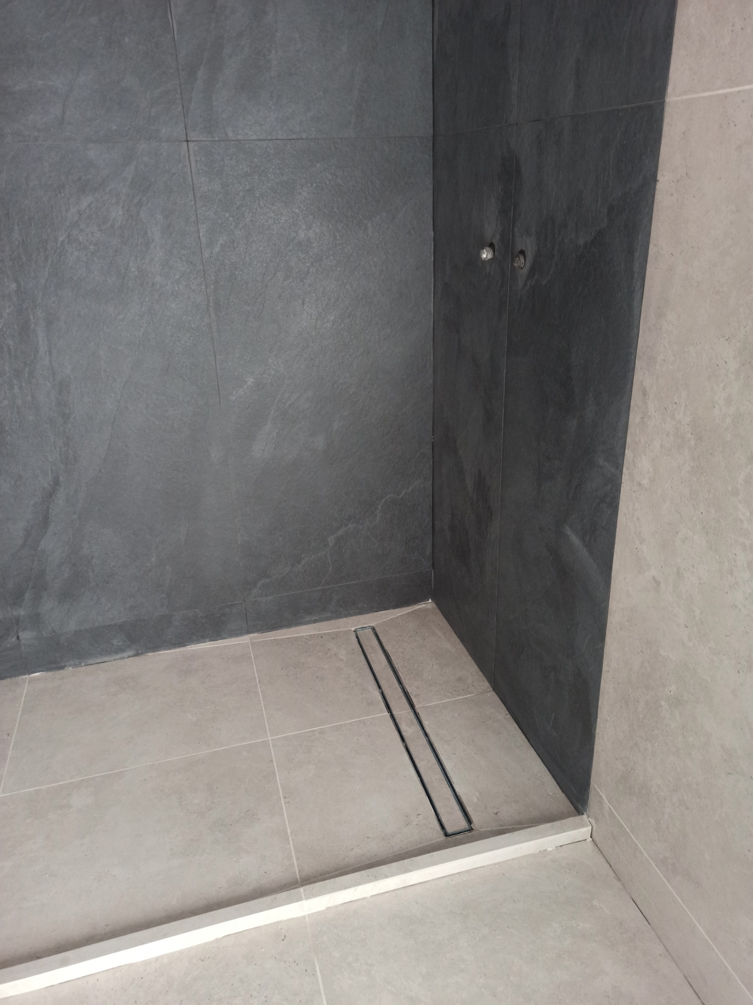 Nowoczesna kabina prysznicowa z szarymi płytkami imitującymi kamień na ścianach i jasnymi płytkami na podłodze. Widoczny odpływ liniowy i minimalistyczne wykończenie.