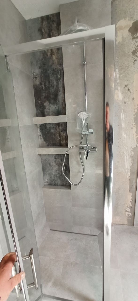 Nowoczesna kabina prysznicowa z szarymi płytkami i wnękami z ciemniejszym akcentem. Chromowana armatura i szklane drzwi. Widoczna dłoń otwierająca kabinę.