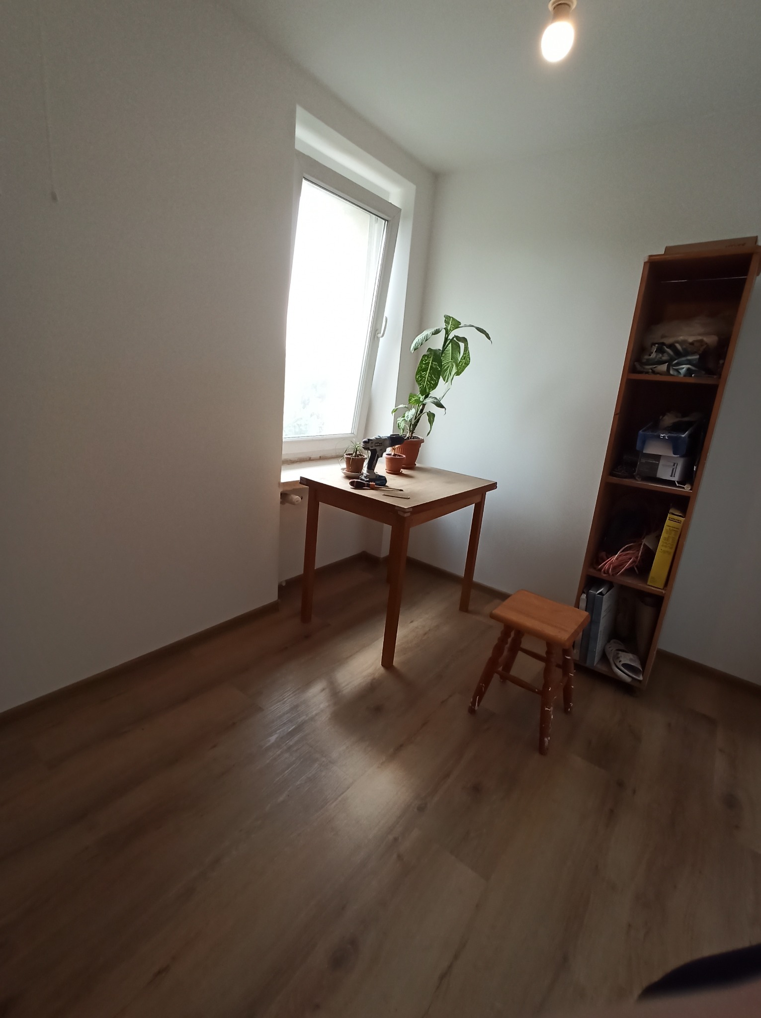 Pokój z drewnianą podłogą, stołem przy oknie, rośliną doniczkową i narzędziami. Widoczny regał z półkami i mały stołek. Przygotowania do pracy.