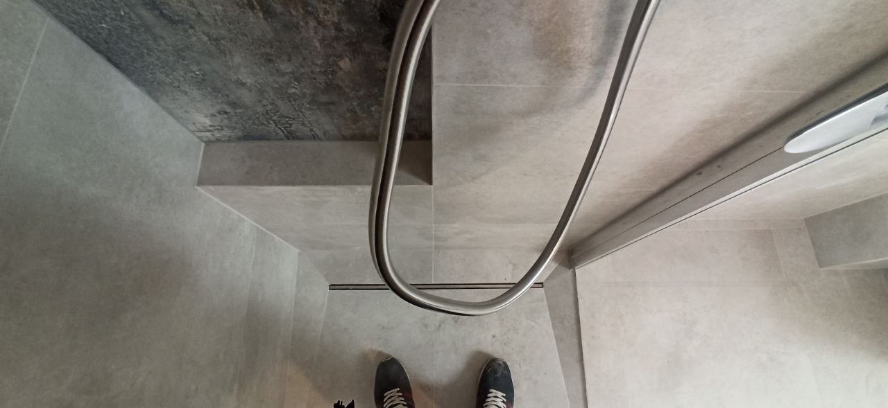 Nowoczesna kabina prysznicowa z odpływem liniowym i szklanymi drzwiami, wykończona szarą płytką imitującą beton. Widoczne stopy osoby fotografującej.