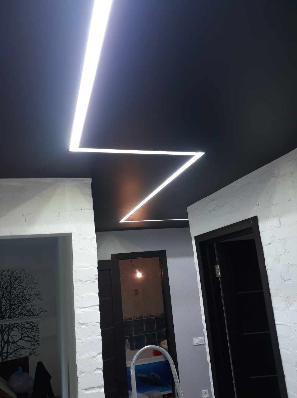 Czarny sufit napinany z geometrycznym oświetleniem LED w kształcie zygzaka, widok z dołu na odnowione wnętrze z białymi ścianami i ciemnymi drzwiami.