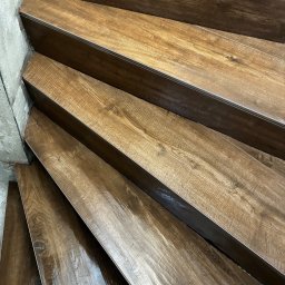 Baubarti - Wykończone schody z płytek imitujących drewno, zabezpieczone metalowymi listwami krawędziowymi. Widoczny fragment ściany z surowego betonu.