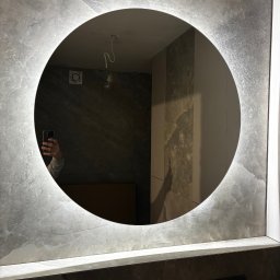 Baubarti - Okrągłe lustro z podświetleniem LED w nowoczesnej łazience wykończonej płytkami imitującymi kamień. Widoczne odbicie osoby robiącej zdjęcie telefonem.