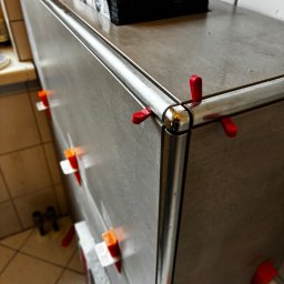 Baubarti - Wykończona obudowa w łazience z szarych płytek, zabezpieczona chromowaną listwą i czerwonymi klinami poziomującymi. Na górze poziomica i paczka płytek.