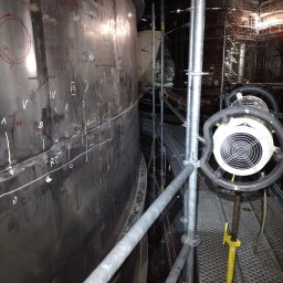 NDTEST - Metalowa konstrukcja zbiornika z oznaczeniami spawalniczymi, oświetlona lampą na rusztowaniu. W tle widoczne rusztowania i elementy konstrukcyjne.