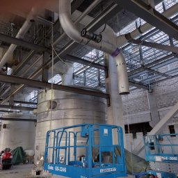 NDTEST - Wnętrze hali przemysłowej z widocznymi rurami, konstrukcjami stalowymi i zbiornikami. Dwie podnośniki koszowe w kolorze niebieskim. Elementy instalacji z oznaczeniami.