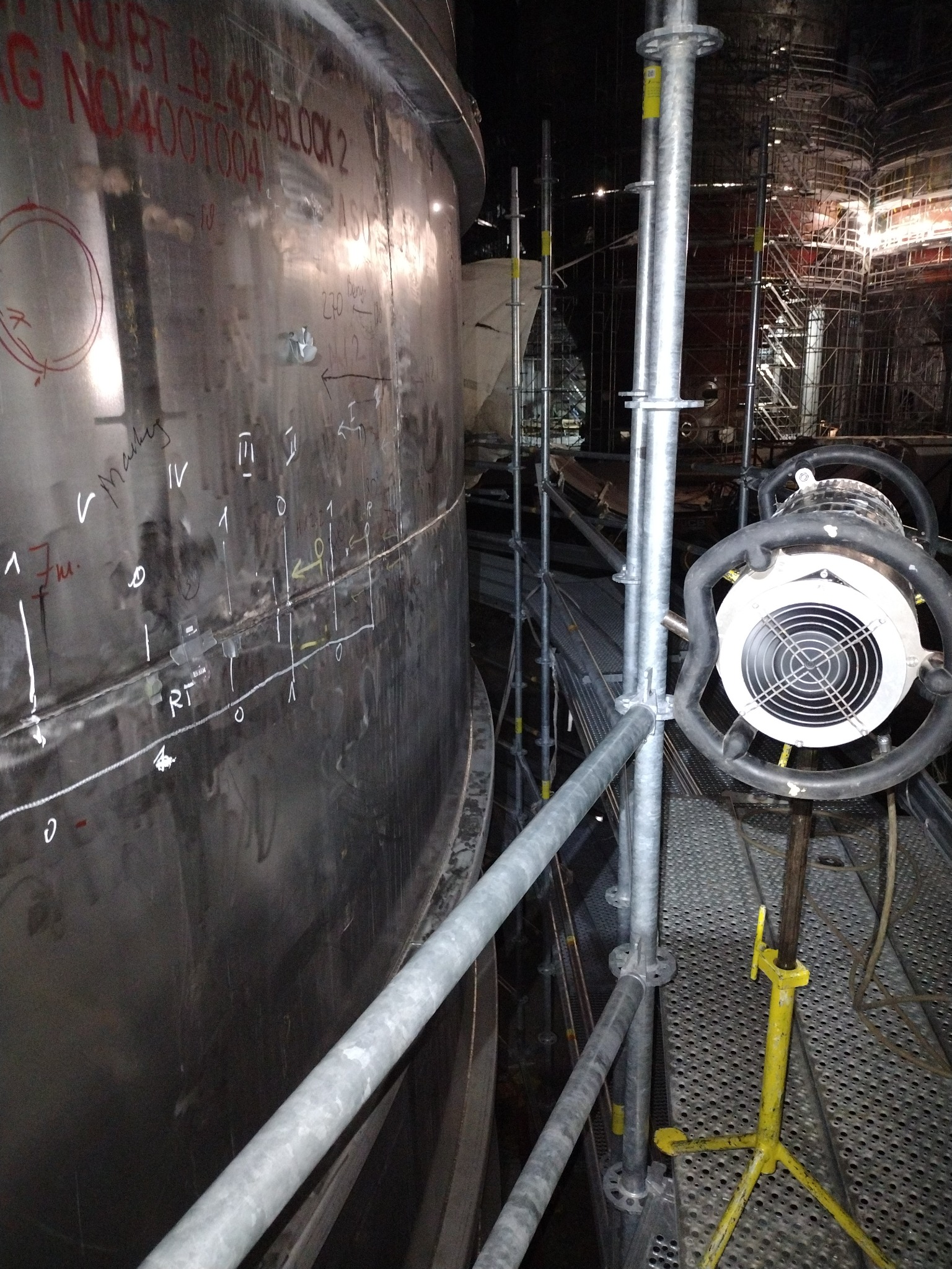 Metalowa konstrukcja zbiornika z oznaczeniami spawalniczymi, oświetlona lampą na rusztowaniu. W tle widoczne rusztowania i elementy konstrukcyjne.