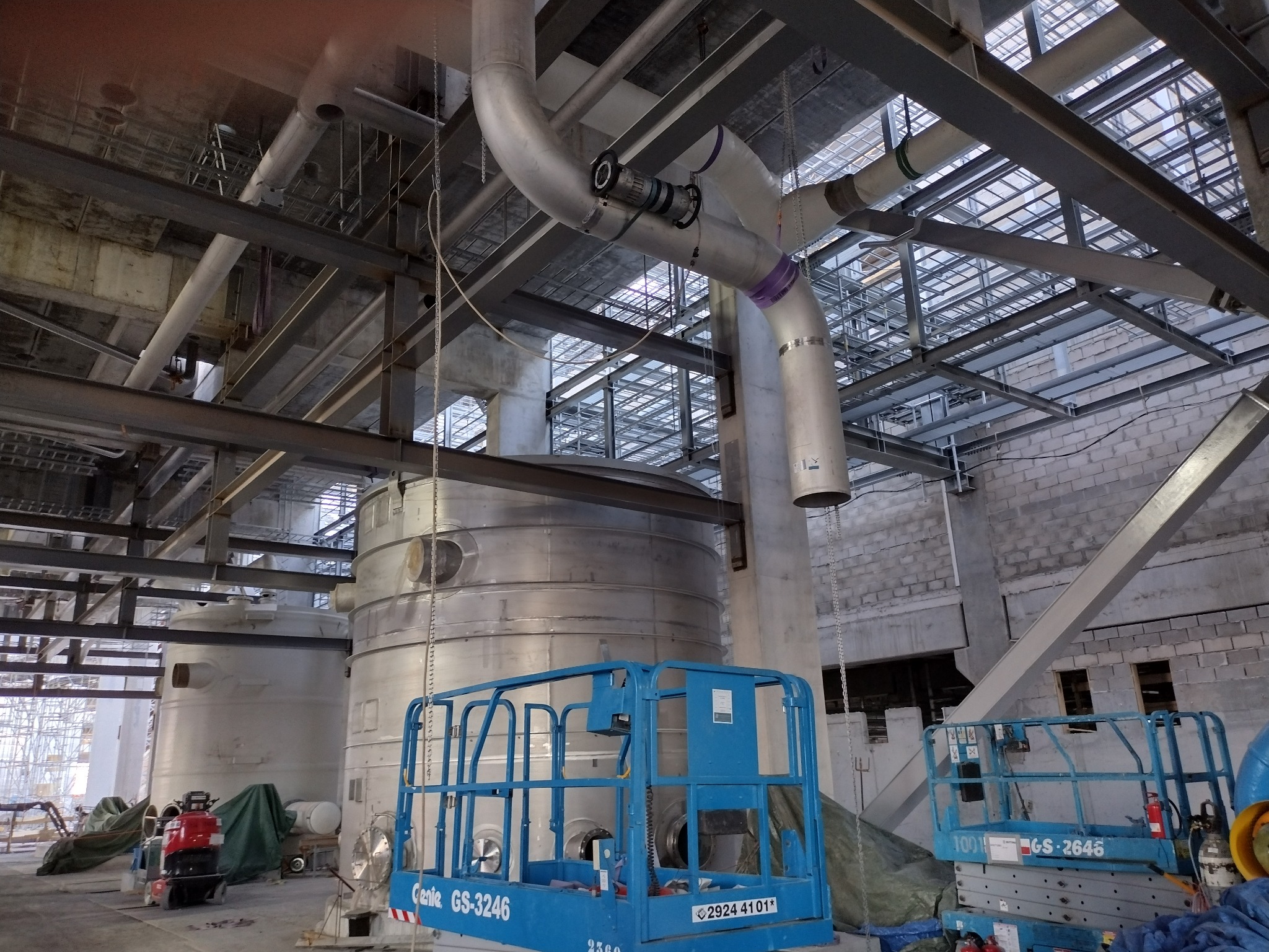 Wnętrze hali przemysłowej z widocznymi rurami, konstrukcjami stalowymi i zbiornikami. Dwie podnośniki koszowe w kolorze niebieskim. Elementy instalacji z oznaczeniami.