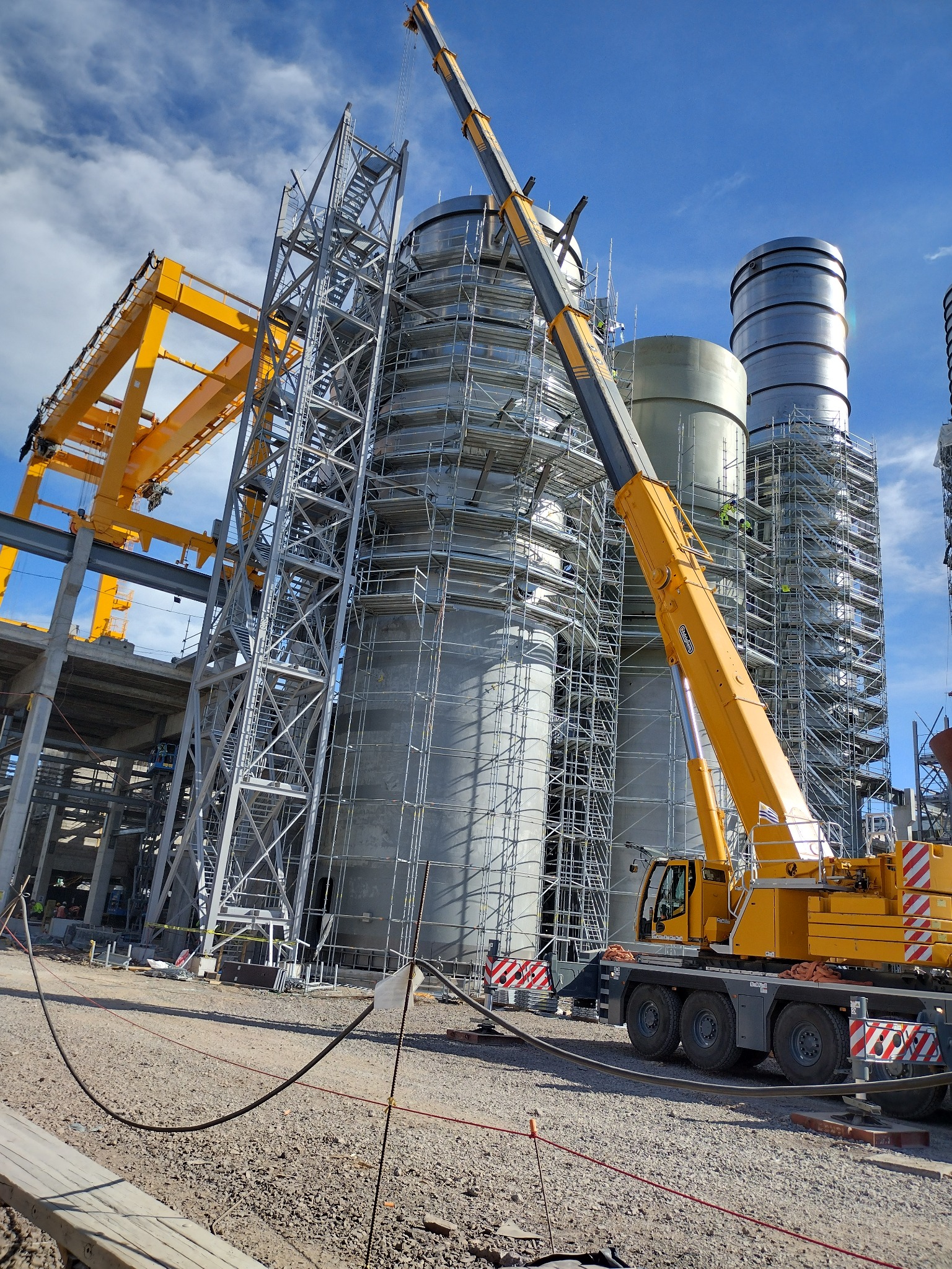 Konstrukcja stalowa w trakcie budowy z użyciem dźwigu, widoczne rusztowania i elementy spawane. Błękitne niebo w tle. Prace na wysokościach.