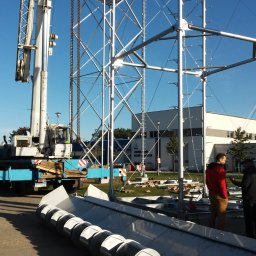 Konsnord Kamil Burblis - Montaż modułowej konstrukcji stalowej z użyciem dźwigu na tle błękitnego nieba i budynku. Widoczne elementy konstrukcyjne i osoby nadzorujące prace.