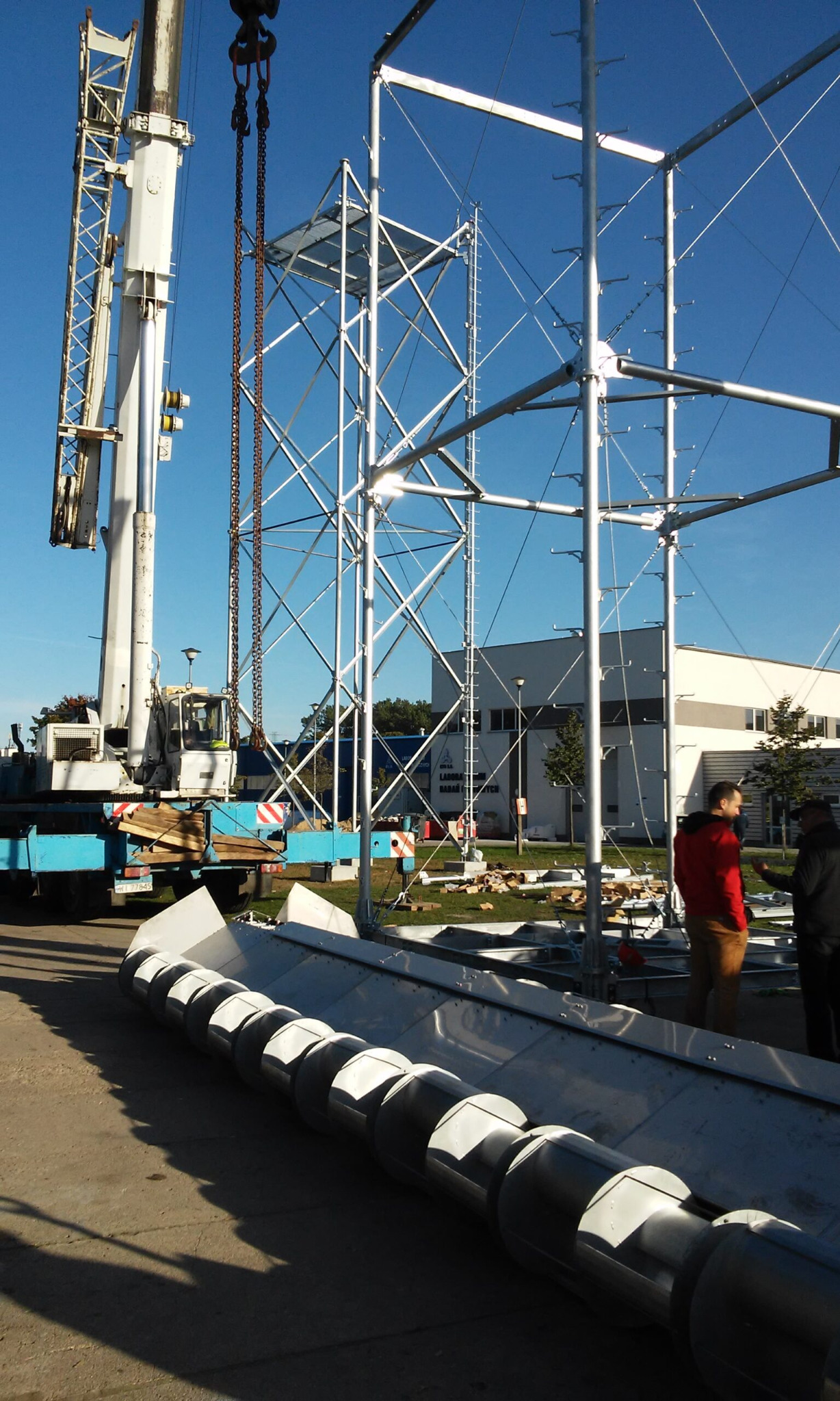 Montaż modułowej konstrukcji stalowej z użyciem dźwigu na tle błękitnego nieba i budynku. Widoczne elementy konstrukcyjne i osoby nadzorujące prace.