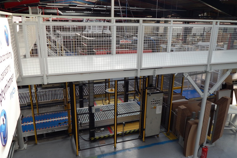 Wnętrze hali produkcyjnej z konstrukcją stalową antresoli i systemem transportu rolkowego. Widoczne elementy sterowania i składowane materiały.