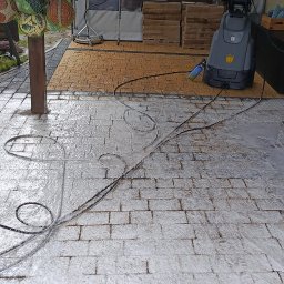 ALTURA - Częściowo umyta kostka brukowa pod zadaszeniem, z widocznym urządzeniem wysokociśnieniowym i stosem kostki. W tle dekoracje i oświetlenie.