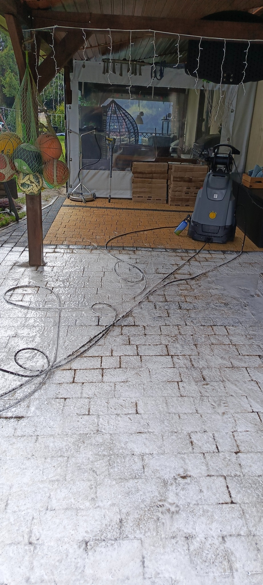 Częściowo umyta kostka brukowa pod zadaszeniem, z widocznym urządzeniem wysokociśnieniowym i stosem kostki. W tle dekoracje i oświetlenie.