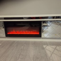 Jakub Zajac - Biała szafka RTV z lustrzanymi frontami i elektrycznym kominkiem z efektem płomieni, na tle szarej ściany i podłogi z paneli. Nowoczesny design.