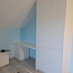 Jakub Zajac - Biała szafa wnękowa z biurkiem pod skosem dachu, widoczne narzędzia na podłodze. Minimalistyczny design, jasne wnętrze, niebieska ściana w tle.