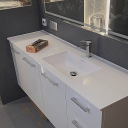 Jakub Zajac - Biała szafka łazienkowa z umywalką i chromowaną armaturą, uzupełniona o pudełko chusteczek. W tle szara ściana z lustrem z podświetleniem LED.